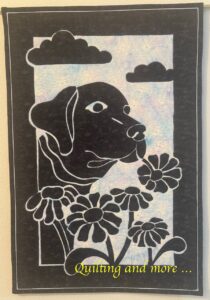Black appliqué dog head on light background. Dog is holding a flower stem in his mouth and has other flowers in t he foreground.