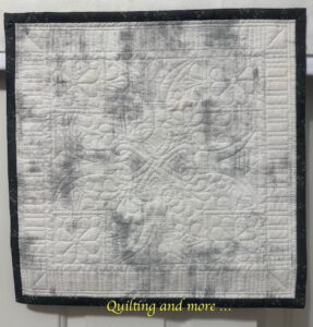 Small (17" square) wholecloth quilt on grey/white Grunge fabric with taupe binding. The quilting is in white thread and is a geometric design repeated four times and surrounded by piano-key quilting in the border.