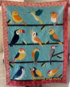 Quilt with brightly multi-colored birds, each different, perching on brown lines of sashing with occasional green leaves. The border is two shades of red done in an attic-window setting to mimic seeing the birds from a window.